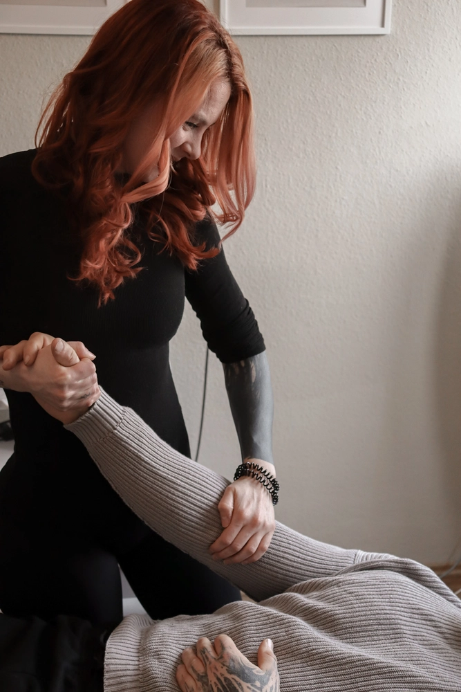 LPZ-Hübner - Physiotherapie und Lymphdrainage - Eine Frau mit langen, gewellten roten Haaren und einem schwarzen Langarmshirt dehnt den Arm einer liegenden Person.