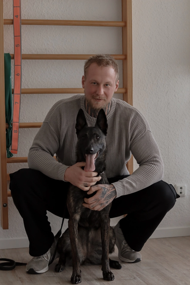 LPZ-Hübner - Physiotherapie und Lymphdrainage - Ein Mann mit Tätowierungen an Händen und Hals hockt und lächelt freundlich, während er einen gestromten Holländischen Schäferhund hält, der die Zunge herausstreckt. Hinter ihnen befindet sich eine hölzerne Sprossenwand und eine weiße Wand.