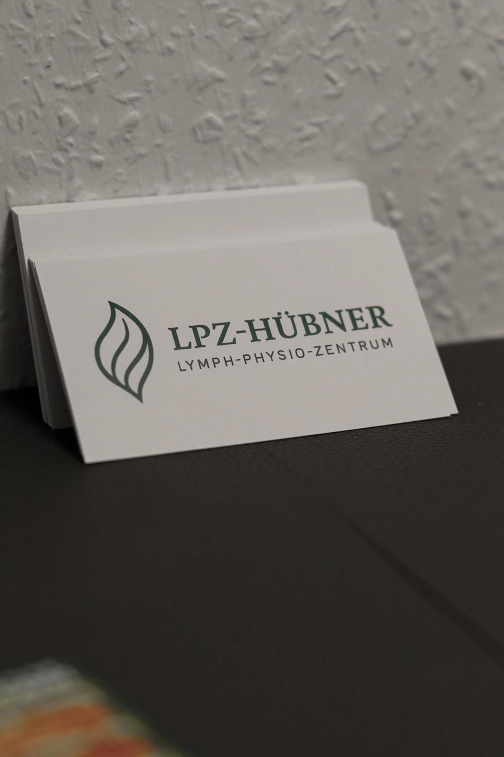 LPZ-Hübner - Physiotherapie und Lymphdrainage - Ein Stapel weißer Visitenkarten für das LPZ-HÜBNER Lymph-Physio-Zentrum mit einem grünen, blattähnlichen Logo und Text lehnt an einer strukturierten Wand.