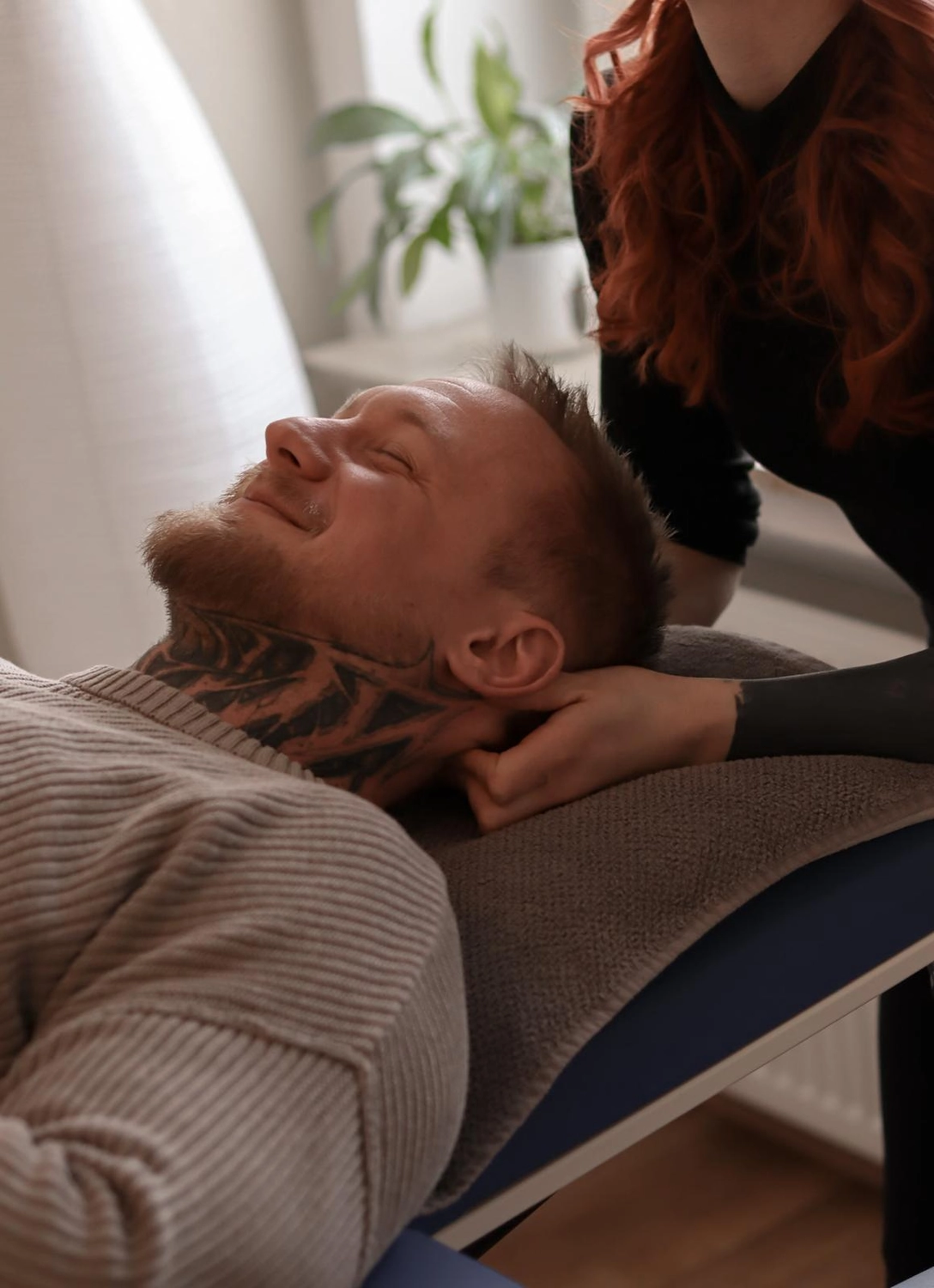 LPZ-Hübner - Physiotherapie und Lymphdrainage - Ein Mann mit einem Nackentattoo liegt mit geschlossenen Augen auf einer Massageliege, während die Hände eines Therapeuten seinen Kopf während einer Behandlung stützen.