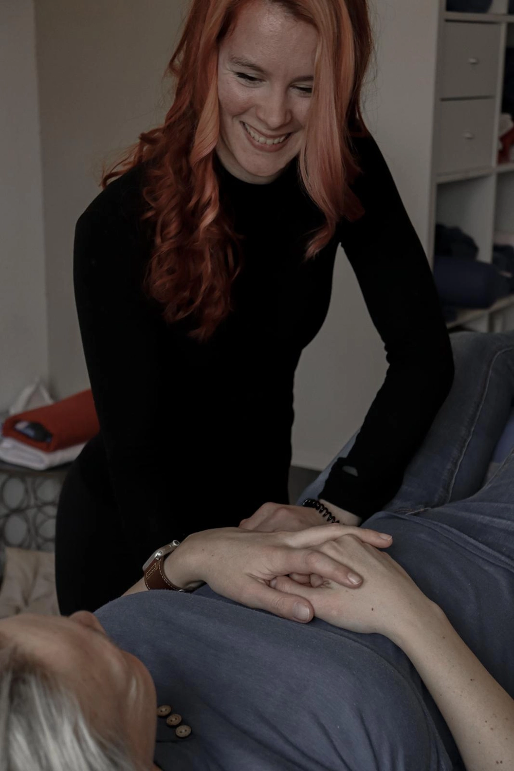 LPZ-Hübner - Physiotherapie und Lymphdrainage - Eine lächelnde Frau mit langen, gewellten roten Haaren beugt sich über eine liegende Person und legt ihr sanft die Hände auf den Bauch – eine wohltuende oder unterstützende Geste.